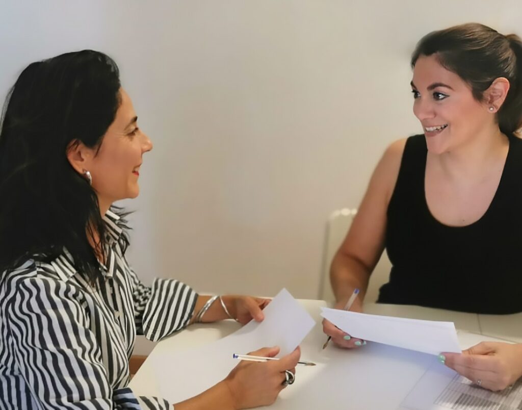 pilar sentada en mesa frente clienta sonriendo las dos y con papeles en mano