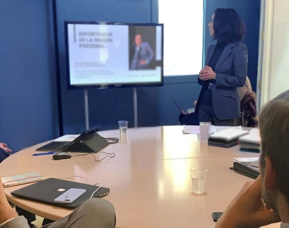 Pilar haciendo jornada empresarial de coaching de equipos en despacho con mesa redonda de madera sentada la gente y ella de pie mostrando cosas en la pantalla frontal